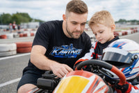Thumbnail for Father wearing Competition Karting shirt helping young driver prepare go kart at karting track